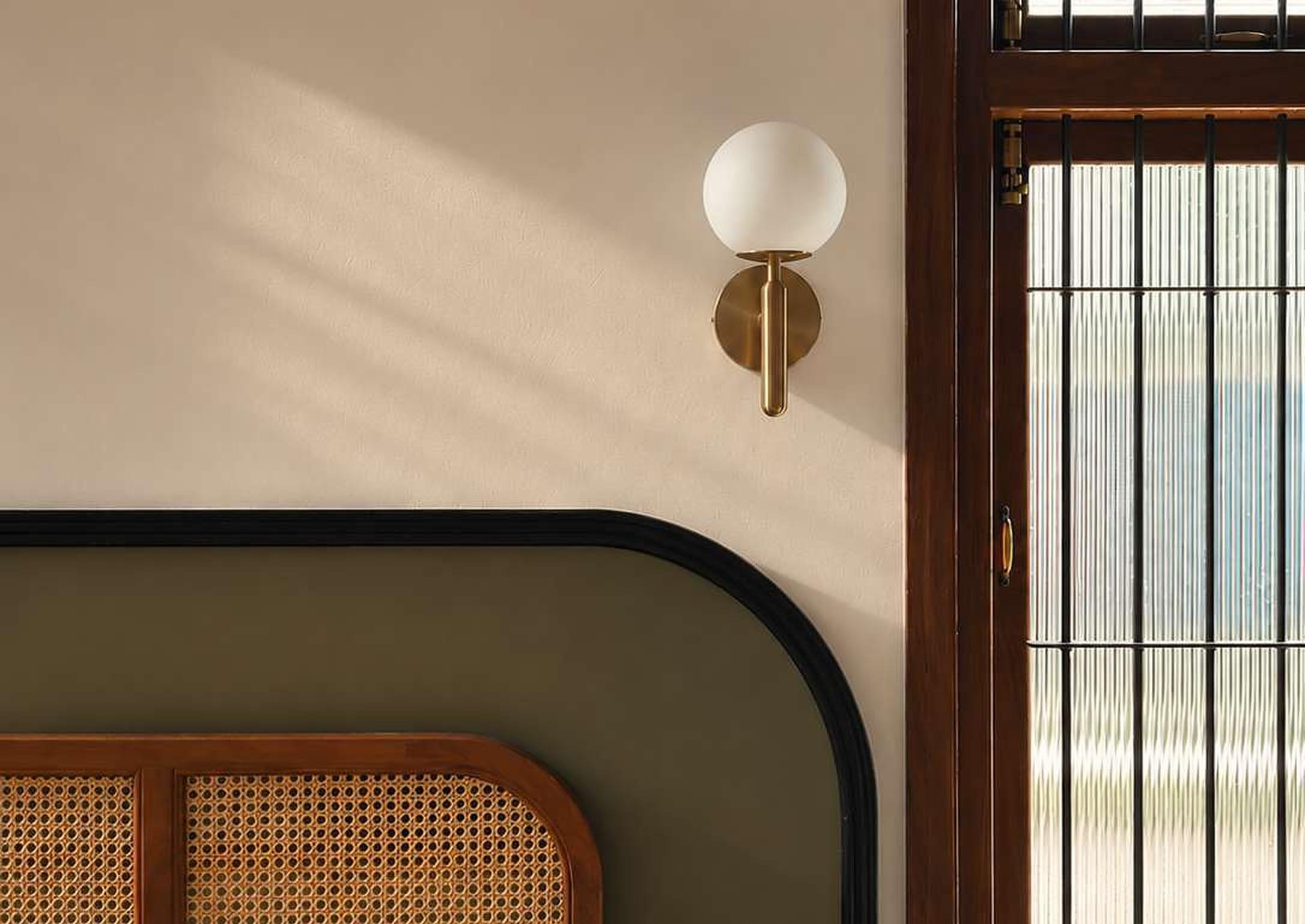 A cane-and-teak headboard with a rounded black trim beneath a brass globe sconce, beside a fluted-glass window framed in dark teak