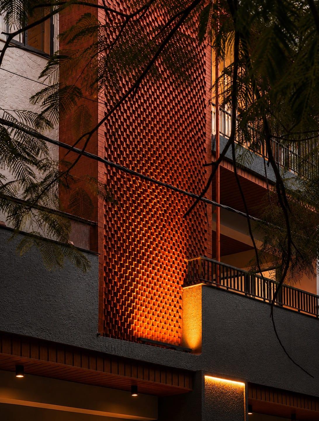 Evening view of the façade: a terracotta jaali screen glowing with uplight beside dark textured plaster and timber balconies