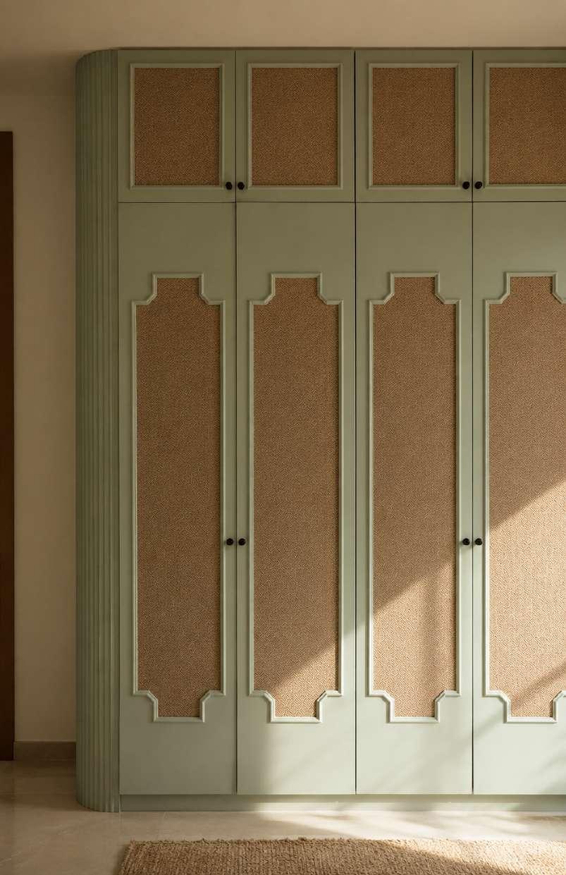 Sage-green wardrobe doors with woven cane panels and a fluted curved edge, lit by low morning sun