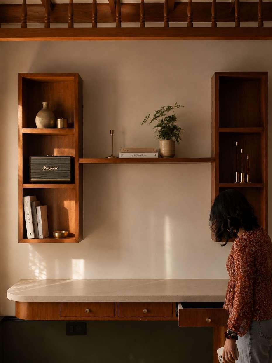 A home workspace with floating teak shelves, a curved stone-topped desk and warm afternoon light raking across the wall